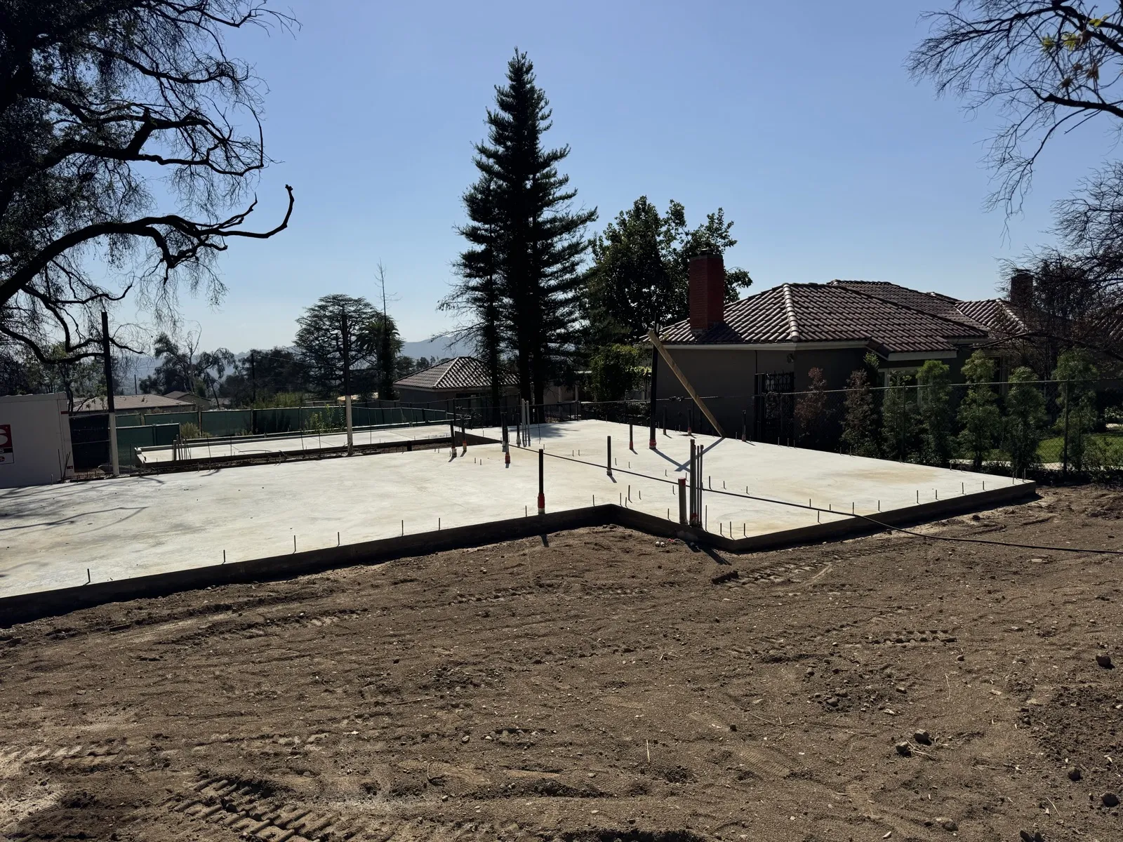 New concrete foundation slab with red-capped plumbing stub-outs and rebar at the East Las Flores Drive fire rebuild, PODS container and neighboring tile-roof homes behind