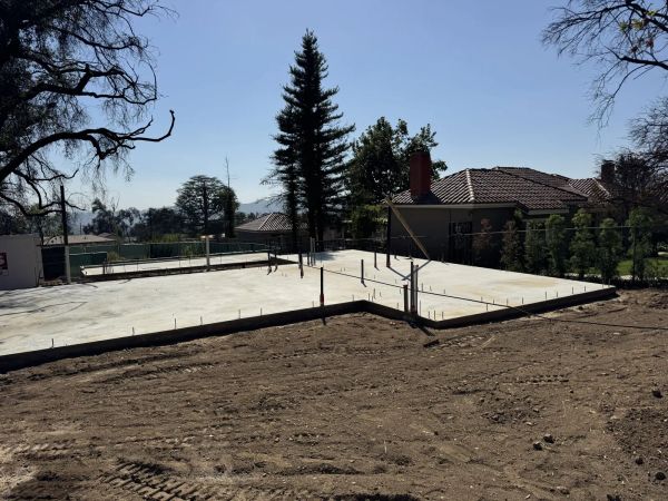 New concrete foundation slab with red-capped plumbing stub-outs and rebar at the East Las Flores Drive fire rebuild, PODS container and neighboring tile-roof homes behind