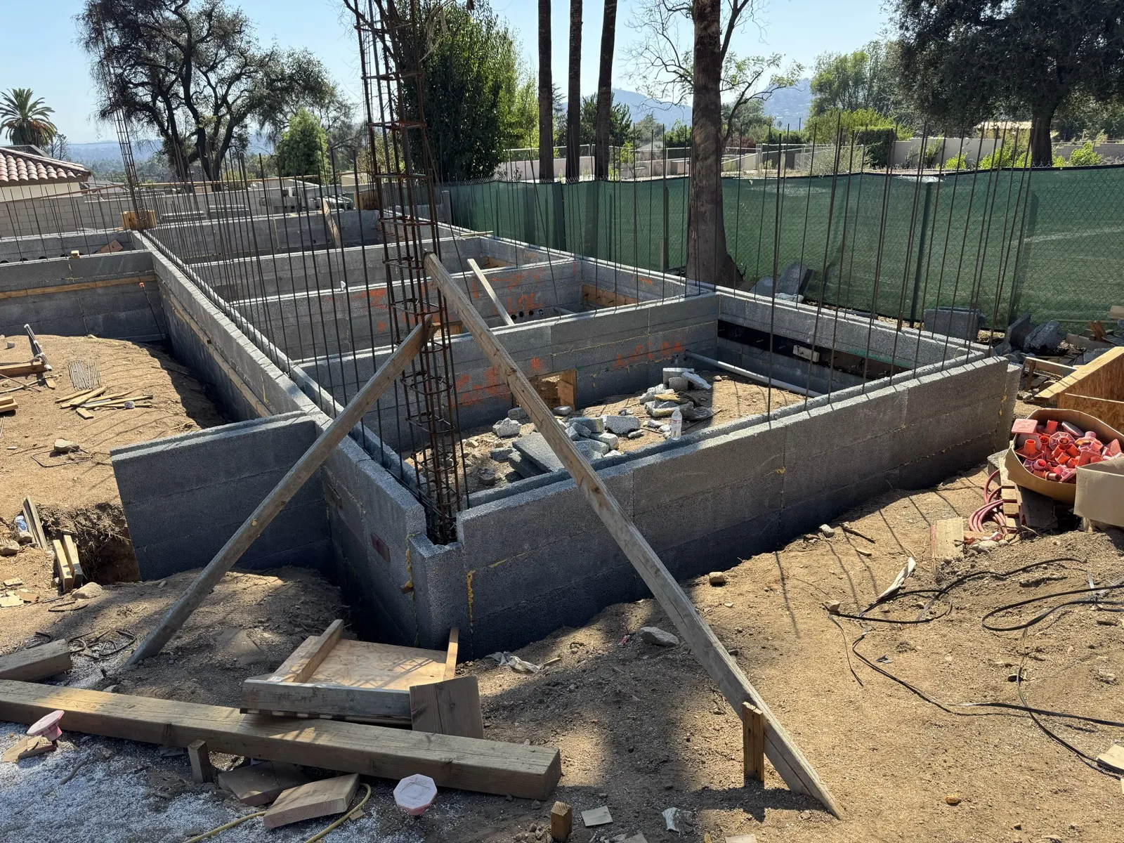Concrete block CMU stem walls forming the house footprint with vertical rebar at the East Palm Street Altadena fire rebuild, wooden form bracing and green mesh fence with oaks and San Gabriel Mountains behind
