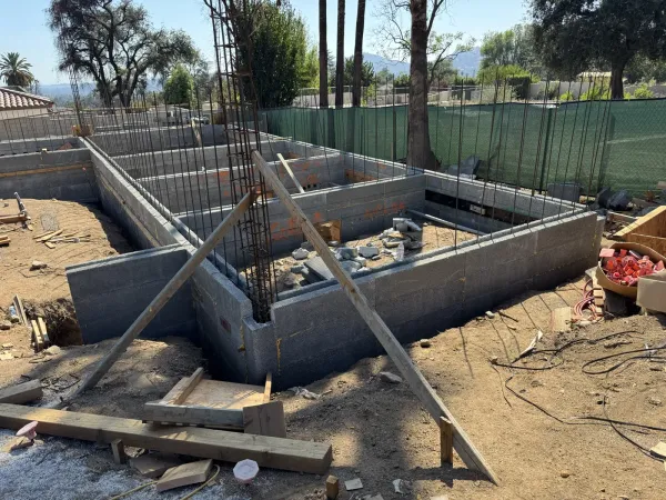 Concrete block CMU stem walls forming the house footprint with vertical rebar at the East Palm Street Altadena fire rebuild, wooden form bracing and green mesh fence with oaks and San Gabriel Mountains behind.