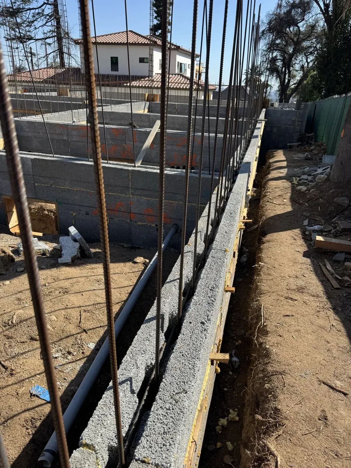 Close view along the top of an ICCF foundation wall with vertical rebar cells at the East Palm Street hillside fire rebuild, excavated grade trench and tile-roof home beyond
