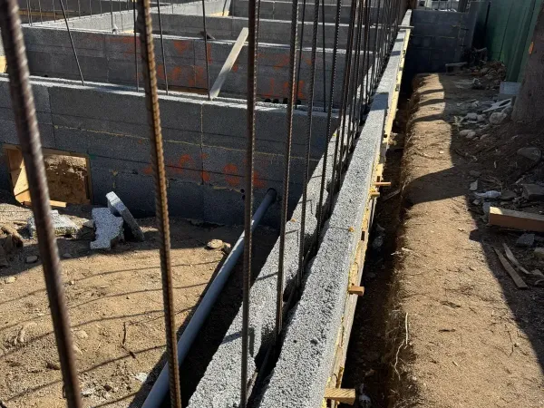 Close view along the top of a CMU retaining wall with vertical rebar cells and PVC drain pipe stubbed through at the East Palm Street hillside fire rebuild, excavated grade trench and tile-roof home beyond.