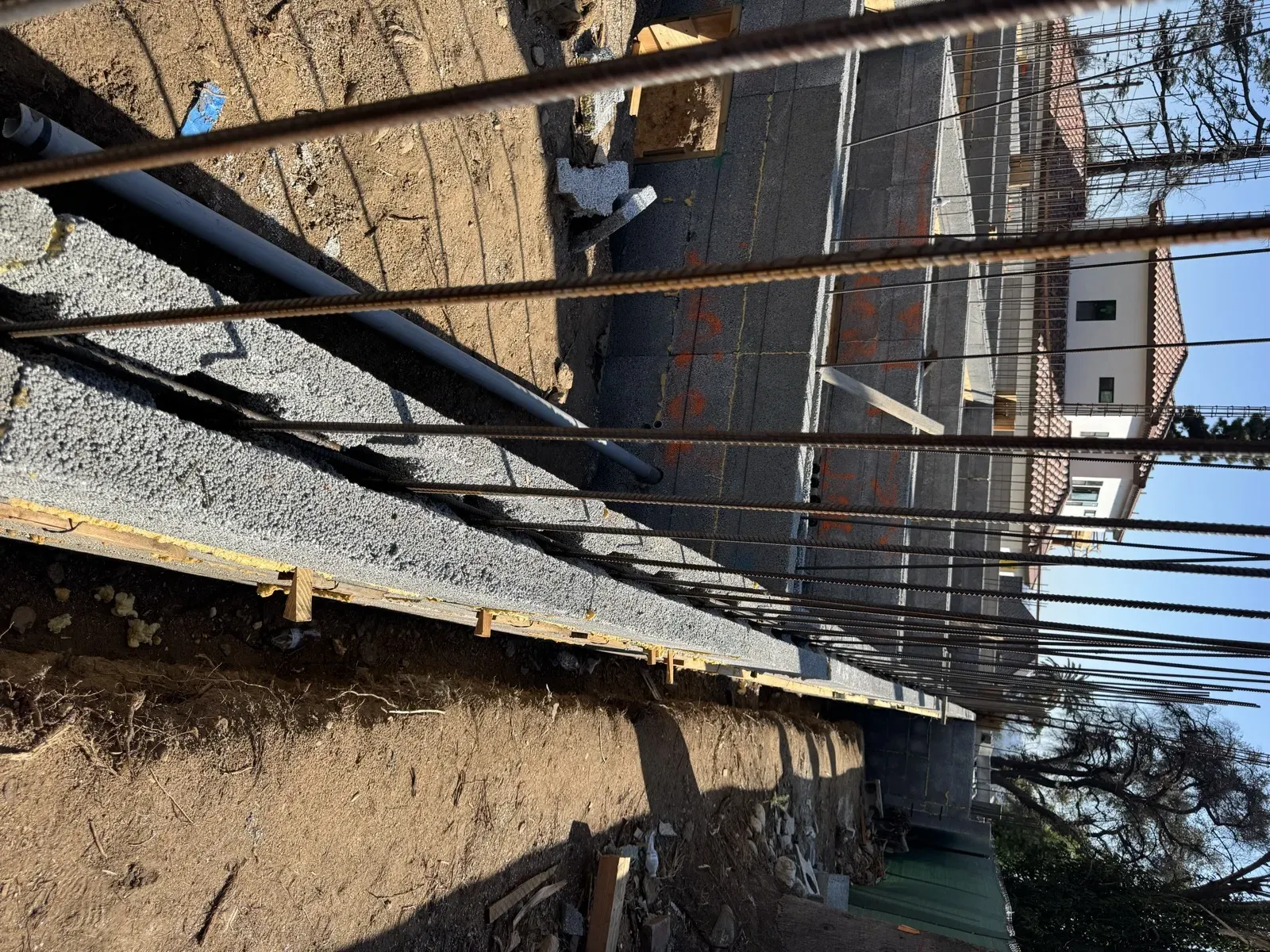 Close view along the top of a CMU retaining wall with vertical rebar cells and PVC drain pipe stubbed through at the East Palm Street hillside fire rebuild, excavated grade trench and tile-roof home beyond