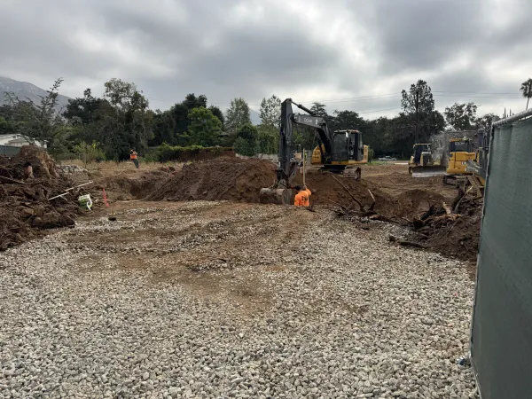 Cellar excavation underway at the Ganesha Avenue Altadena multi-unit fire rebuild with John Deere excavator and crew working in the dig next to the crushed rock construction entrance
