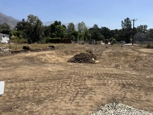Cleared and graded building pad on a hillside lot showing rough earthwork with tire tracks from grading equipment, ready for footing excavation on the Altadena fire rebuild site. Orange staking flags mark property boundaries and drainage flow lines across the compacted dirt.