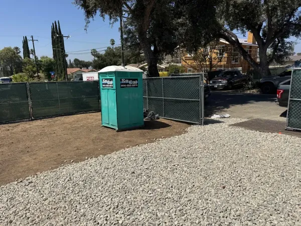 Temporary construction entrance with crushed rock base and green privacy fence along the property line for the multi-unit rebuild. Portable toilet and site security fencing indicate active construction phase on the Ganesha Avenue lot.