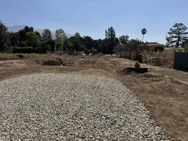 Compacted gravel base prepped for driveway and approach slab at the front of the fire rebuild lot. Rough grading extends toward the rear with minor grading swales directing stormwater runoff around future building pads.