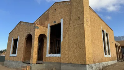 Framed two story fire rebuild house at the Glenrose Avenue Altadena site with OSB sheathing, arched entry, and black window frames set against a blue sky with San Gabriel Mountains