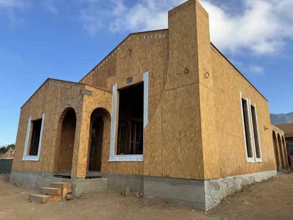 Framed two story fire rebuild house at the Glenrose Avenue Altadena site with OSB sheathing, arched entry, and black window frames set against a blue sky with San Gabriel Mountains