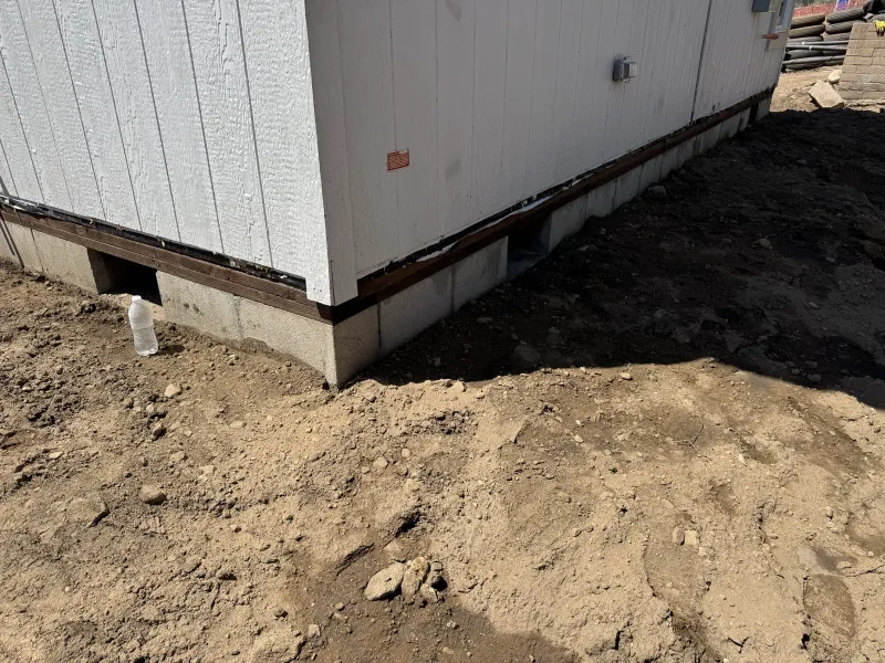 Altadena fire-rebuild hillside lot at the foundation phase with a white-sided building on a concrete base with brown trim, a small red plaque sign on the wall, and freshly graded uneven dirt