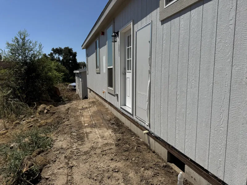 Newly placed gray manufactured home with white trim, white door, and black lantern sconce on a concrete foundation at a Las Flores Drive Altadena fire rebuild, with exposed tracked brown dirt revealing concrete footings and a white PVC pipe, dry native shrubs, a wooden fence, and blue sky