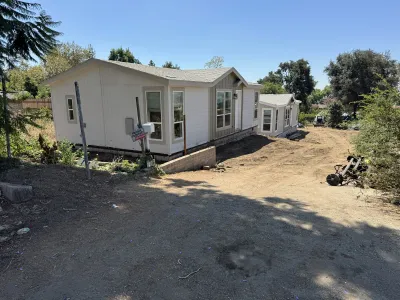 Newly placed white manufactured home on a graded dirt hillside lot in Altadena during an Eaton Fire rebuild, with a low concrete retaining wall, Sigalert construction sign, and a red mini skid steer