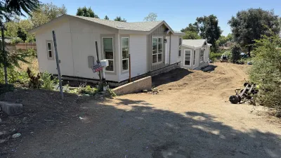 Newly placed white manufactured home on a graded dirt hillside lot in Altadena during an Eaton Fire rebuild, with a low concrete retaining wall, Sigalert construction sign, and a red mini skid steer