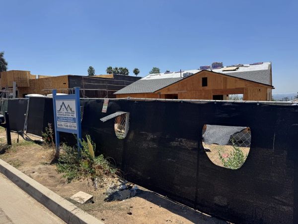 Newly framed Altadena fire-rebuild house on a residential street corner behind a black mesh fence and blue Scott J. Woodland Construction sign, with wooden studs, OSB sheathing, black house wrap, and gray asphalt shingles over roof trusses, plus palm trees and hazy San Gabriel Mountains behind