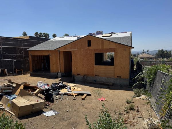 Altadena fire-rebuild house in structural framing phase on a bare dirt lot with a concrete stem wall foundation, OSB plywood sheathing, rough window openings, a partially shingled roof, and a white corrugated water tank in front