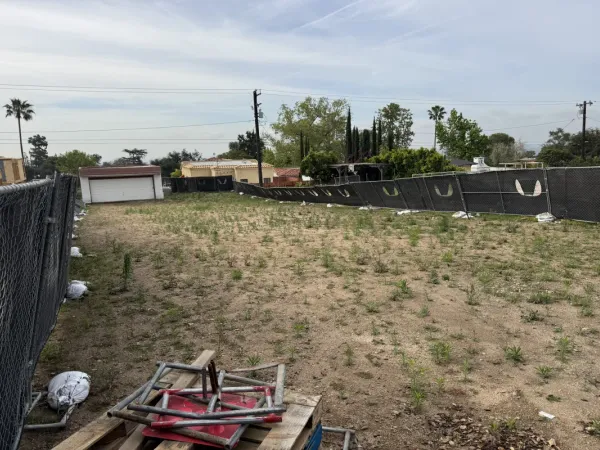 Pre-construction view of Altadena fire rebuild lot showing cleared dirt site with chain-link fencing, privacy screening, and construction materials
