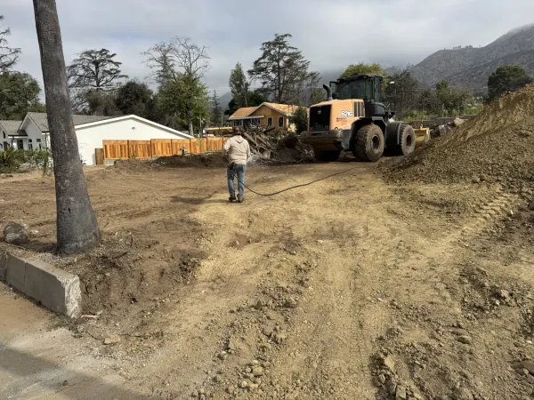 Worker in white sweatshirt holding a hose near a Caterpillar 824C wheel loader on the Altadena Punahou Street fire-rebuild lot during rough grading, with a dirt pile, wooden privacy fence, white house, and cloudy San Gabriel Mountain foothills