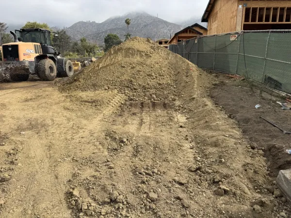 Case 621G wheel loader parked beside a large excavated dirt pile from cut and fill on the Altadena Punahou Street fire-rebuild lot, with a green mesh Custom Fence screen, wood-framed house under construction, and misty San Gabriel Mountains