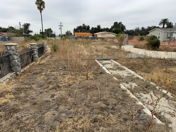Vacant fire rebuild lot in Altadena with remaining concrete foundation strips and adjacent wrought iron fence with stone pillars after Eaton Fire demolition