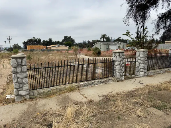 Altadena W Mendocino Ave vacant lot viewed from sidewalk behind existing stone pillar and wrought iron fence, post-Eaton Fire demolition site