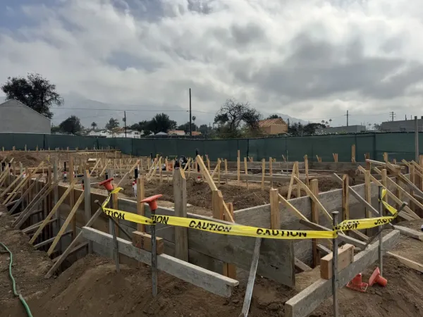 Active construction site featuring excavated foundation trenches and wooden concrete formwork with wooden stakes and boards defining the future building footprint, caution tape and orange rebar safety caps visible
