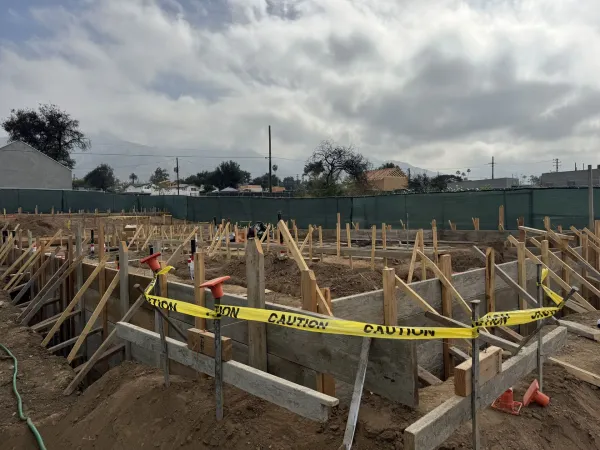 Active construction site featuring excavated foundation trenches and wooden concrete formwork with wooden stakes and boards defining the future building footprint, caution tape and orange rebar safety caps visible