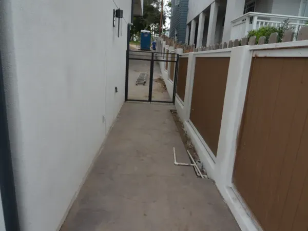 Narrow concrete walkway between two residential buildings showing white stucco exterior, brown paneled fence, PVC pipes, and loose soil during construction