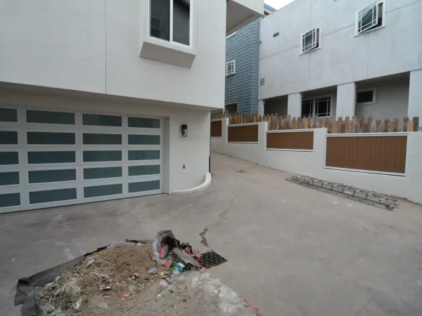 Modern residential construction with white stucco building, frosted glass garage door, concrete driveway, and tiered concrete retaining wall with wood fence