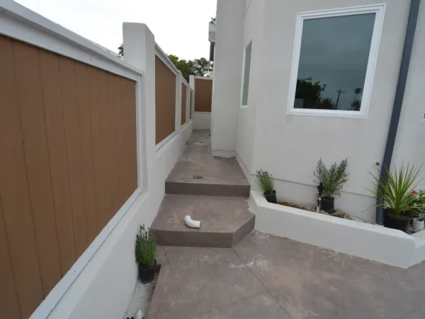 Side yard construction of modern home featuring textured concrete walkway, white stucco walls, perimeter wall with brown vertical board fence, and built-in planters
