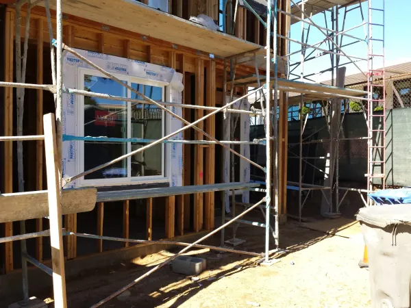 Wood framing and structural details of building under construction with installed white-framed window, MOISTOP flashing, metal scaffolding, and uneven dirt grading