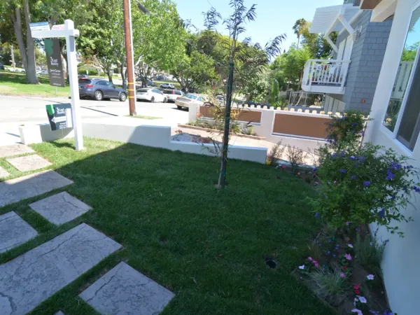 Professionally landscaped residential front yard with large-format textured gray concrete pavers, green lawn, low white stucco retaining wall, and horizontal wood-slat fence