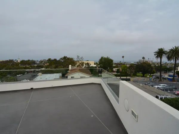 Flat rooftop deck with dark gray waterproof membrane, integrated surface drains, white parapet walls, and frameless glass railing overlooking coastal urban landscape
