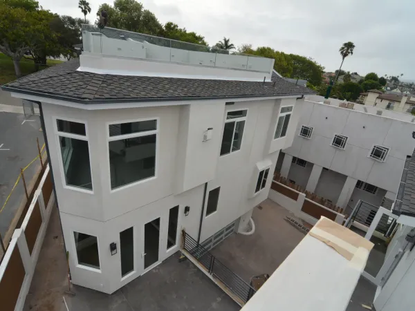 High-angle drone shot of modern residential construction site featuring white stucco home with glass-railed rooftop deck, paved courtyard, and final site grading