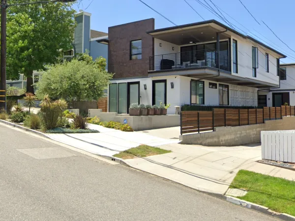 Completed modern hillside residential project on St Lucia Avenue in Redondo Beach featuring two connected 2-story container homes with bridge, white stucco and brown textured panels, cantilevered balcony, concrete retaining walls, and drought-tolerant landscaping