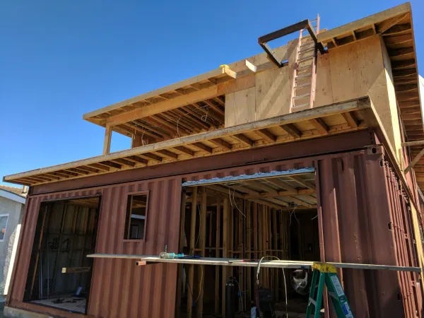 Two-story shipping container shell with corrugated steel walls integrated with wood-frame upper stories and utility rough-in