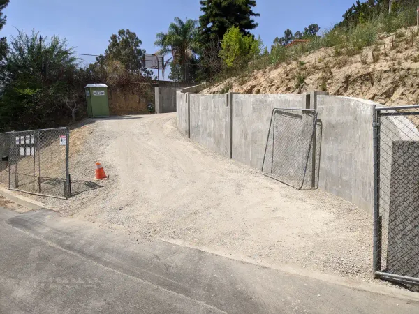 A construction site featuring a newly built, curved concrete retaining wall that runs along a graded dirt and gravel driveway leading up a steep slope
