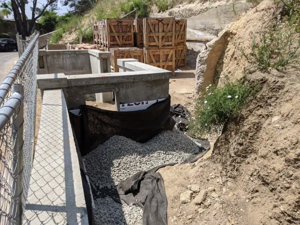 A construction site with new concrete retaining wall and crushed gravel driveway bed lined with black landscape fabric on a steep graded slope