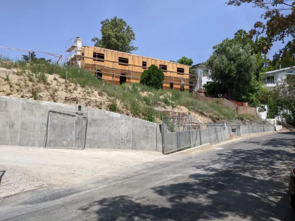 A residential hillside construction project showing a large concrete retaining wall, sloped grading with protective fencing, and a wood-framed house under construction