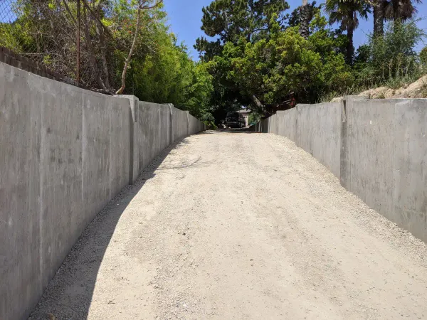A long, sloped gravel driveway under construction bordered by tall poured concrete retaining walls on a residential hillside