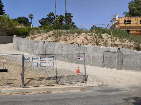 A construction site on a hillside featuring a large, multi-section concrete retaining wall with a dirt and gravel driveway winding up the slope
