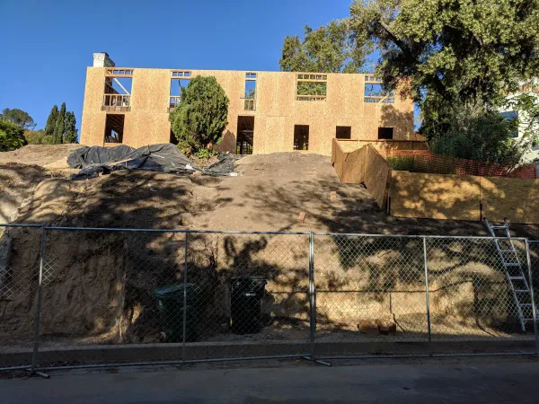 Two-story residential house under construction with OSB sheathing sits atop a steep, newly graded dirt slope with erosion control measures