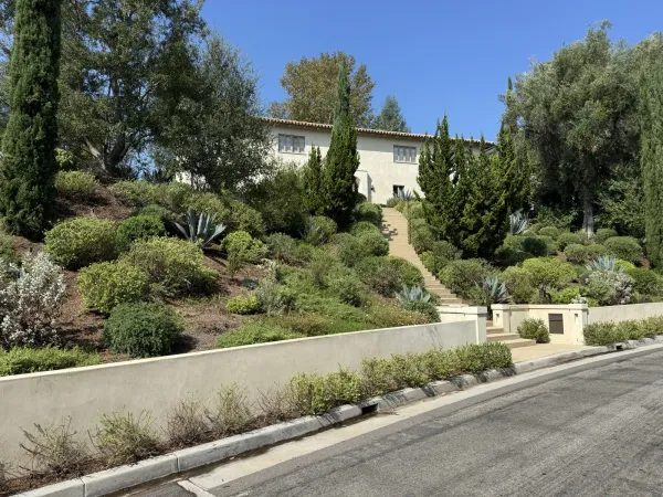 Completed two-story stucco house with a terracotta tile roof behind a concrete retaining wall and stepped entry path on the steep landscaped Redcliff Street hillside