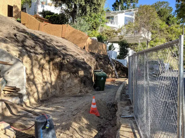 Residential hillside construction site with steep dirt grading supported by plywood retaining walls, driveway entrance from street